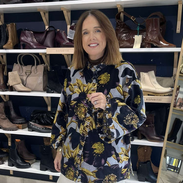 A woman stands in a shoe and boot store wearing a black, gold, and white floral long-sleeve top with white cropped pants and black flats. Shelves behind her display various styles of boots and shoes.