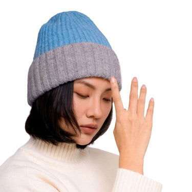 A woman wearing a two-tone ribbed knit beanie hat in light blue and gray, paired with a cream turtleneck sweater against a plain white background.