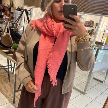 A woman takes a mirror selfie in a boutique, wearing a chunky coral pink fringed scarf, a beige jacket, a black top, and brown wide-leg trousers.