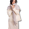 A woman models a light beige knit scarf with fringed edges, wrapped around her shoulders and neck over a cream-colored sweater and skirt, set against a plain white background.