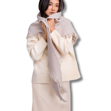 A woman models a light beige knit scarf with fringed edges, wrapped around her shoulders and neck over a cream-colored sweater and skirt, set against a plain white background.
