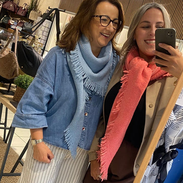 Two women taking a mirror selfie in a clothing boutique. The woman on the left wears a light blue cardigan, a light blue scarf, and striped white pants.