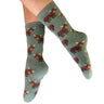 A pair of sage green crew socks with a repeating pattern of fluffy red and brown Highland cows, shown on a person's feet against a plain white background.