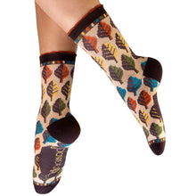A pair of crew socks with a colorful autumn leaf pattern on a beige background, featuring dark brown ribbed cuffs, heels, and toes, shown on a person's feet against a white background.