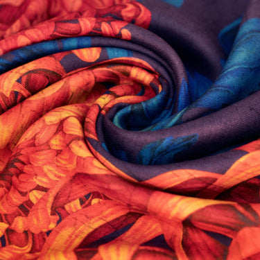 A close-up of a lightweight fabric scarf or textile draped in a spiral, showing a bold floral pattern in orange, red, yellow, and deep blue.