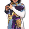 A woman models a light blue scarf with a large purple and yellow floral print, worn wrapped around her shoulders over a white long-sleeve top against a plain white background.