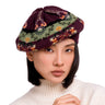 A close-up of a woman wearing a knitted beret hat with a dark burgundy base and a green, orange, and white Nordic-style patterned band, paired with a cream ribbed turtleneck sweater against a solid white background.