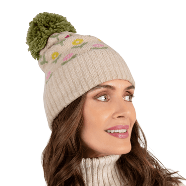 Woman wearing a beige knit beanie with floral patterns and a green pom-pom on a white background