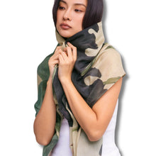 A woman modeling a lightweight scarf with a green, black, and beige camouflage pattern, wrapped around her neck and shoulders against a plain white background.