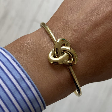 A gold-plated knot cuff bracelet worn on a person's wrist, shown against a light gray wood floor background with a blue and white striped shirt cuff visible.