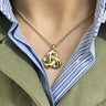 A close-up of a person wearing a gold pendant necklace with an interlocking knot design, styled over a blue and white striped collared shirt and an olive green jacket.