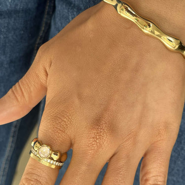 A close-up of a person's hand wearing a gold ring with a round clear gemstone and a gold bracelet with a chunky bamboo-like design. The person is wearing blue jeans and a blue-and-white striped shirt.