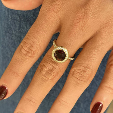 A close-up of a hand wearing a gold ring with a round dark brown gemstone and a halo of small clear stones, shown against blue jeans and a striped shirt cuff.
