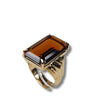 A gold ring featuring a large, rectangular faceted amber-brown gemstone, shown on a plain white background with a soft drop shadow.