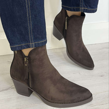 A pair of dark brown suede ankle boots with side zippers, shown on a model wearing dark blue jeans against a light gray floor and white wall.