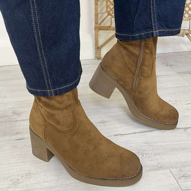 A person wearing brown suede ankle boots with a chunky block heel and elastic side gore, paired with blue jeans, standing on a light wood floor.