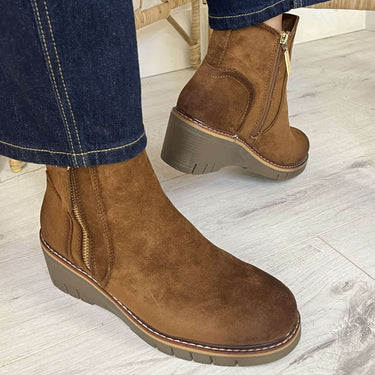 A person wearing brown suede ankle boots with dark gray wedge heels and blue jeans, standing on a light wood floor.