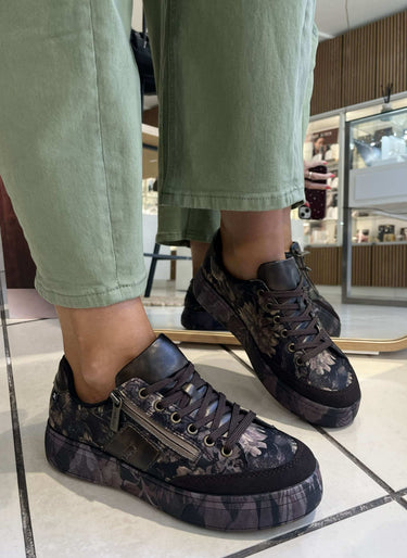 A person wearing dark brown and black floral lace-up sneakers with olive green pants, standing on a tiled floor inside a store.