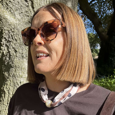 Woman wearing sunglasses and a patterned scarf, standing next to a tree outdoors.