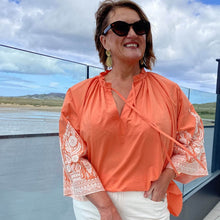 Woman wearing an orange blouse with lace sleeves standing outdoors with a scenic background.
