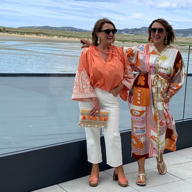 Two women standing by a waterfront, one wearing an orange blouse and white pants, the other in a patterned dress.