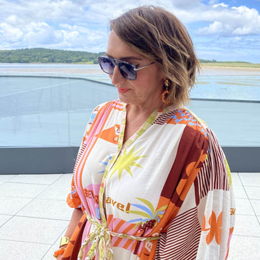 Woman wearing a colorful kimono-style garment with sunglasses, standing by a waterfront.
