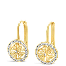 A pair of gold huggie hoop earrings with circular medallion pendants featuring a floral cutout design, surrounded by a halo of small clear stones, set against a plain white background.