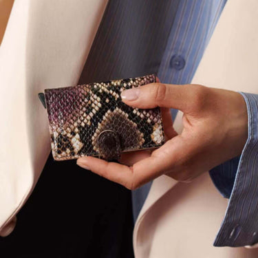 A close-up of a hand holding a small snakeskin-pattern wallet or card holder against a softly blurred background of clothing.