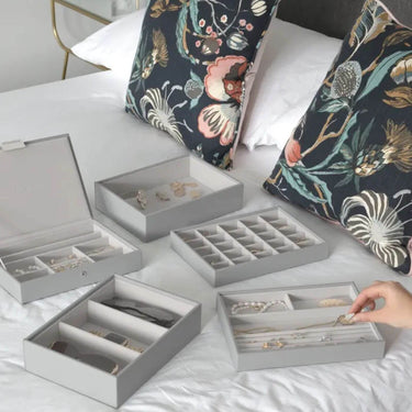 A set of light gray jewelry storage boxes arranged on a white bed, with a hand placing jewelry into one box. The boxes include different compartments and are shown with various pieces of jewelry inside.
