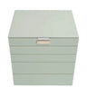 A square jewelry box with a hinged lid and five horizontal drawers, finished in a pale sage green color with a small beige brand plaque on the front.