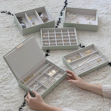 Four light sage green jewelry boxes with multiple compartments, shown open on a white textured rug. Hands are visible holding one of the boxes, and the boxes contain various pieces of jewelry including rings, necklaces, bracelets, and earrings.