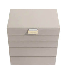 A square jewelry box in a light taupe-beige faux leather finish with a small gold-tone metal accent on the front, shown on a plain white background.