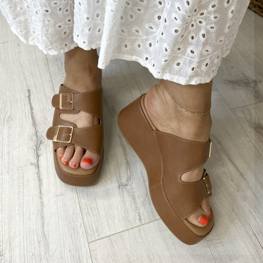 Brown sandals worn with a white lace dress on a light wooden floor.