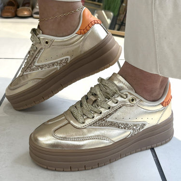 gold sneakers with brown soles worn indoors on a tiled floor.
