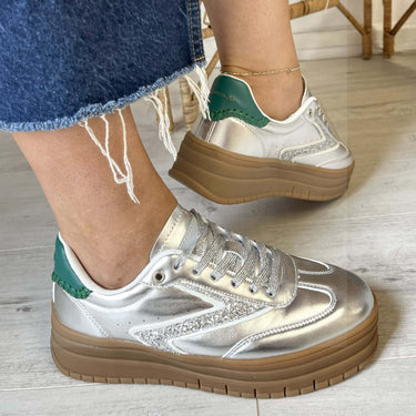 Silver sneakers with green accents worn on a light wooden floor.