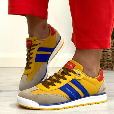 Yellow and beige sneakers with blue accents worn with red pants on a light wooden floor.
