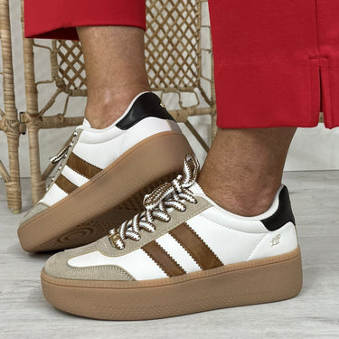 A person wearing red trousers and brown-and-white platform sneakers with three stripes, standing on a light wood floor in front of a woven chair.
