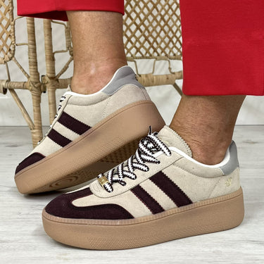 A person wearing beige platform sneakers with dark burgundy suede panels and three dark stripes, paired with bright red pants, standing on a light wood floor.