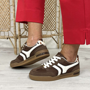A person wearing brown and white low-top sneakers with red pants, standing on a light gray floor in front of a woven rattan screen.
