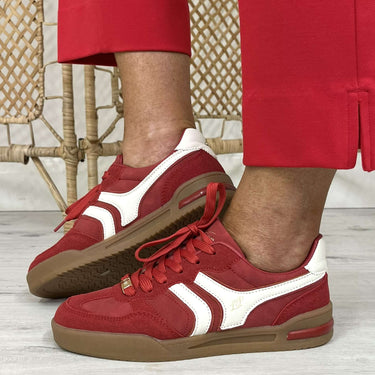 Red and white low-top sneakers worn with red trousers, shown from the knees down on a light wood floor in front of a woven cane screen.