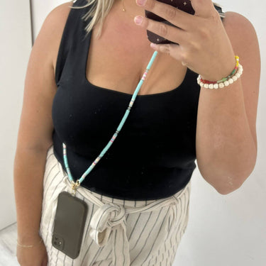 A person taking a mirror selfie wearing a turquoise and pink beaded phone strap with a dark gray phone attached, paired with a black tank top and cream pinstriped pants.