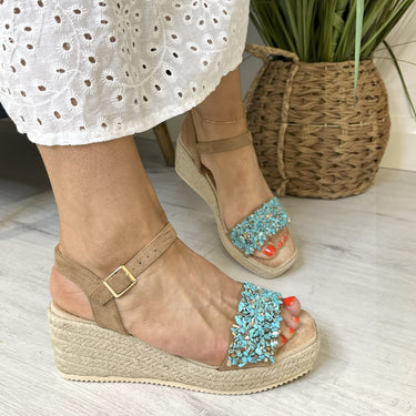 Beige wedge sandals with turquoise embellishments worn indoors.