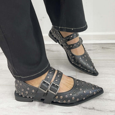 Black studded shoes with buckles worn with black pants on a light wooden floor.