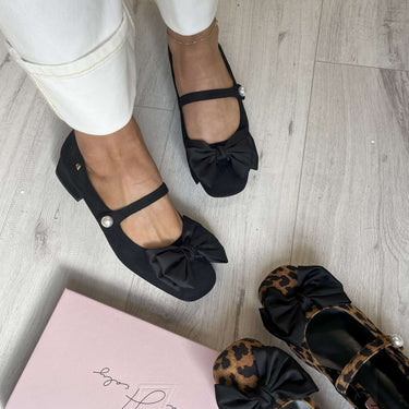 Black flats with bow details on a light wooden floor, next to a pink box.