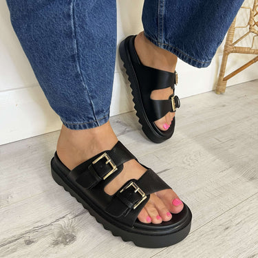 Black platform sandals with gold buckles worn with blue jeans on a light wooden floor.