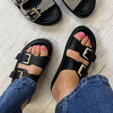 Black sandals with gold buckles worn on a light wooden floor.