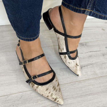 Pointed-toe shoes with black straps worn with blue jeans on a light wooden floor.
