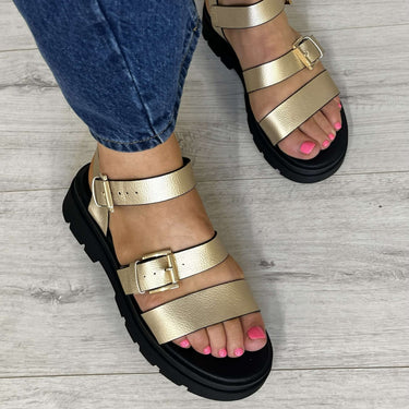 Gold sandals worn with blue jeans on a light wooden floor.