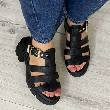 Black strappy sandals worn with blue jeans on a light wooden floor.