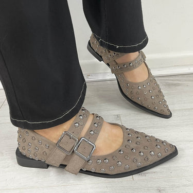 Brown studded flats with buckles worn with black pants on a light wooden floor.